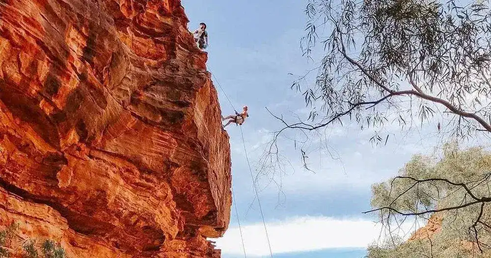 Unusual Things To Do in Kalbarri 15 This image shows two people abseiling off a steep, rust-colored rock face in a natural, rugged environment. One climber is near the top of the cliff while the other is suspended in mid-air, descending with ropes, surrounded by trees and a blue sky in the background. The scene evokes a sense of adventure and exploration.