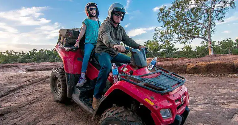 Unusual Things To Do in Kalbarri 16 This image shows two people riding a red quad bike on a dirt trail, likely during an outdoor tour or adventure. The driver is wearing a helmet and sunglasses, while the passenger is enthusiastically smiling with her mouth open, wearing a helmet and seated behind. The rugged terrain and trees in the background suggest they are in a natural, off-road environment, enjoying an exhilarating quad bike experience. Water bottles are attached to the front of the bike, indicating they are prepared for a longer ride.