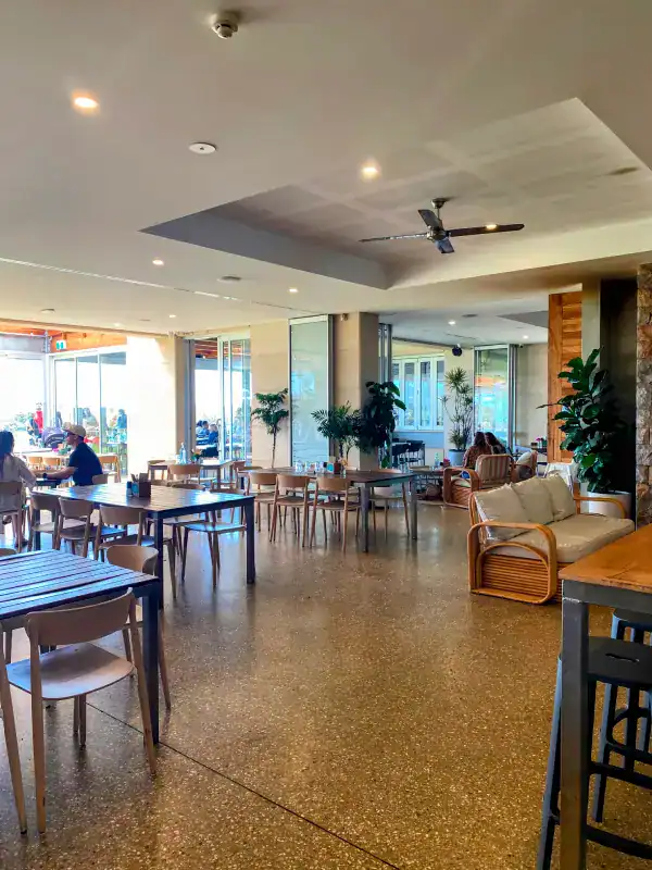 The interior of Eagle Bay Brewing Co in Western Australia, featuring an open and airy design with modern wooden tables and chairs, potted plants, and large windows letting in natural light. The space includes a relaxed seating area with wicker furniture and cushions.