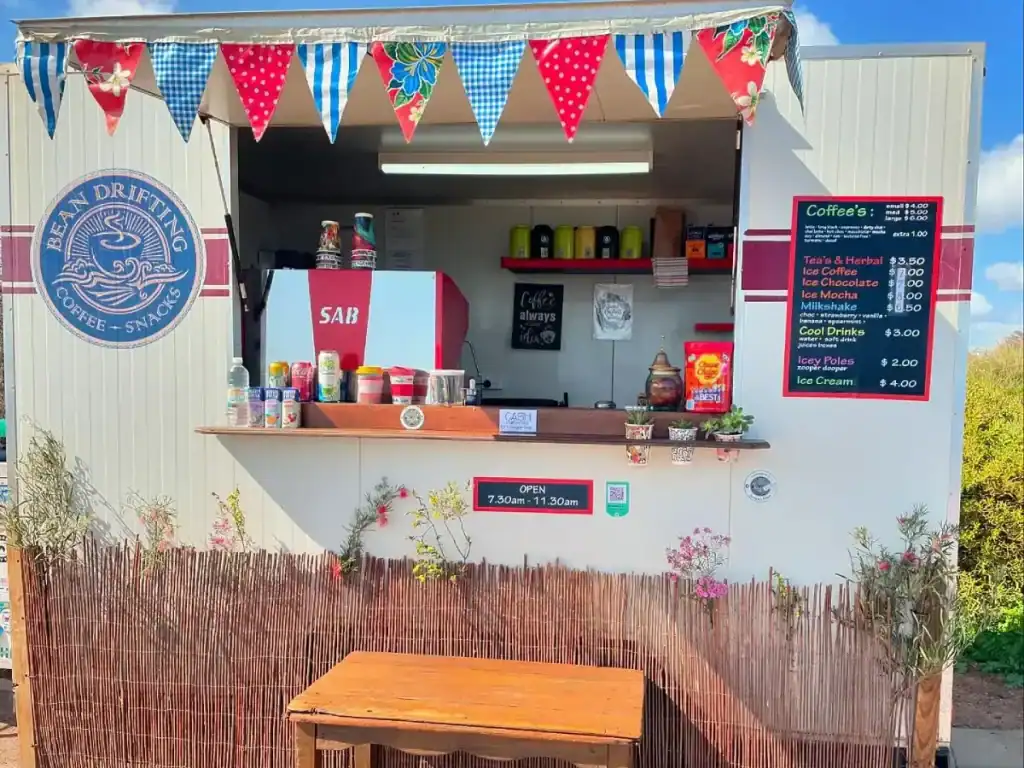 The Best Kalbarri Restaurants, Bars, and Cafes: My Guide for 2026 19 Bean Drifting coffee stand in Kalbarri, featuring a small, decorated counter with colorful bunting, a large coffee machine, and a menu board listing various coffee drinks and snacks. The stand is surrounded by plants and offers drinks like iced coffee, milkshakes, and herbal tea, with hours posted as 7:30 AM to 11:30 AM